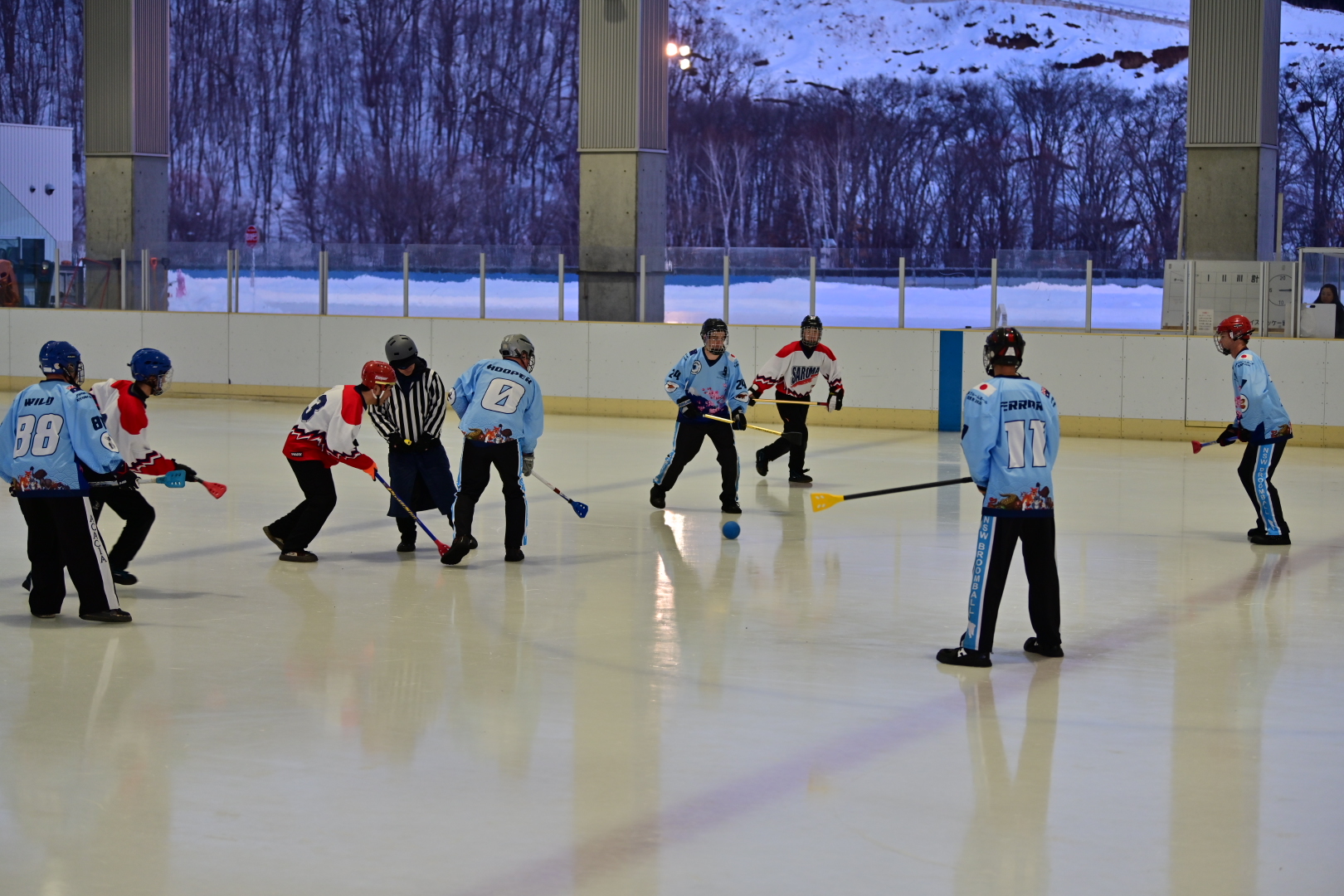 Players face off on an outdoor ice rink with snow-covered hills behind